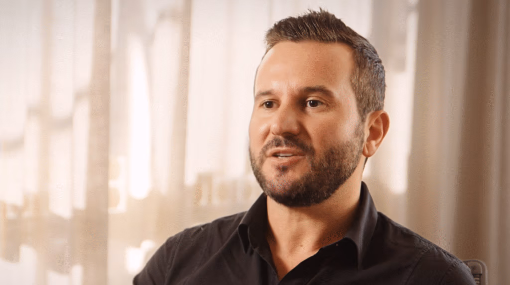 A man with neatly styled short brown hair and a trimmed beard is speaking. He is wearing a black button-up shirt. The background is softly lit with beige curtains and blurred light, creating a warm, neutral setting.