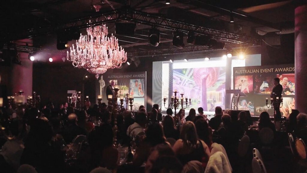 A large crowd seated at round tables watches a brightly lit stage at the Australian Event Awards. A presenter stands at a lectern; colourful screens and banners are visible. A grand chandelier hangs above the elegant, darkened venue.
