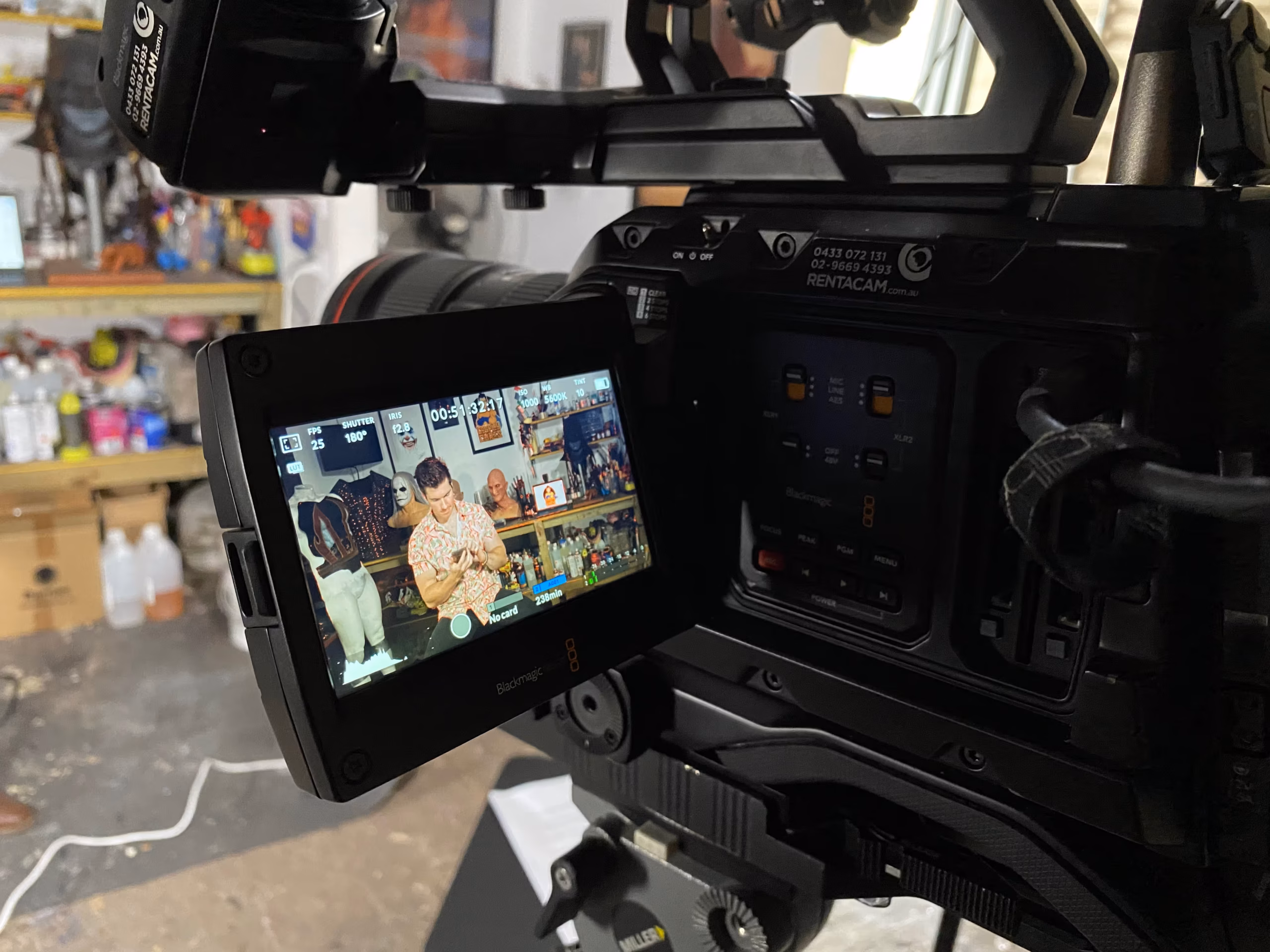 Close-up of a professional video camera used for video production Sydney, its display screen showing two people working at a table with drinks and snacks in a colourful, decorated room. The background is slightly blurred, with the camera dominating the right side.