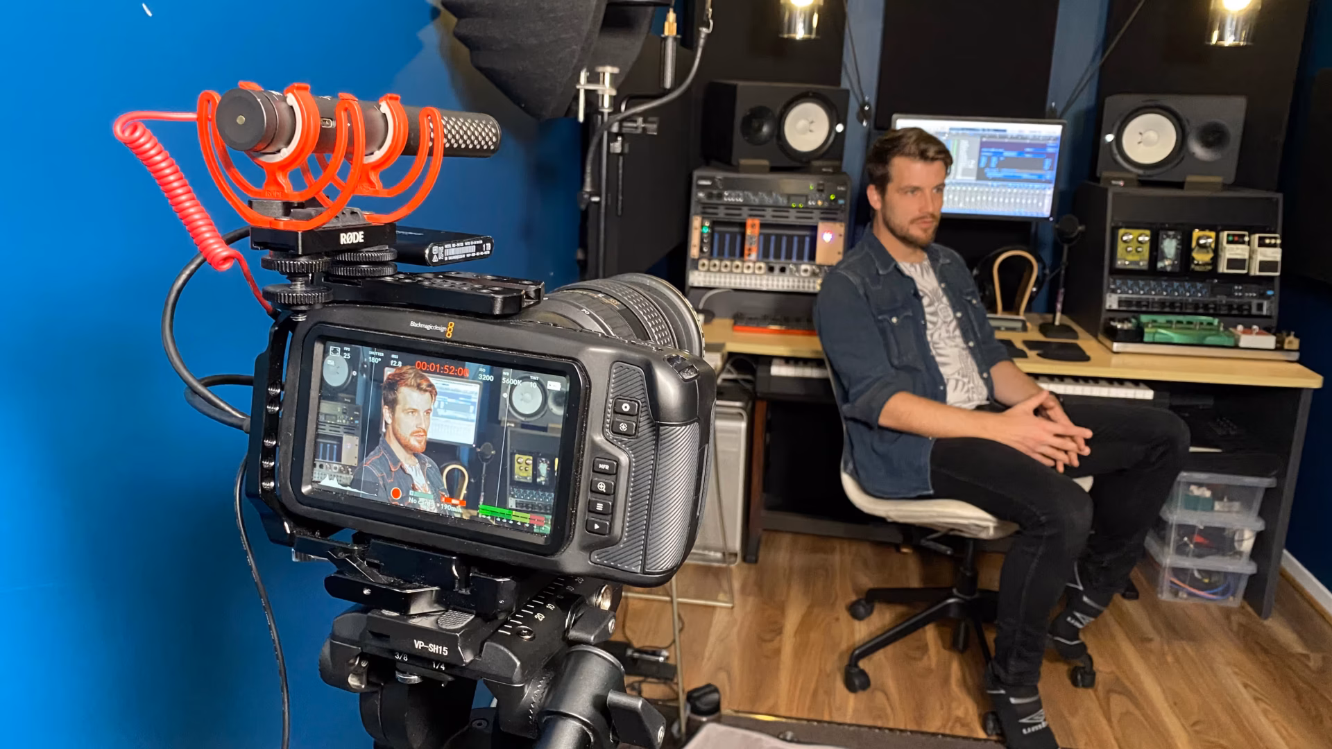 A man sits on a chair in a home recording studio, facing the camera, surrounded by audio equipment and computer screens. Studio lighting and a microphone are set up nearby, capturing his image for video production Sydney professionals would appreciate.