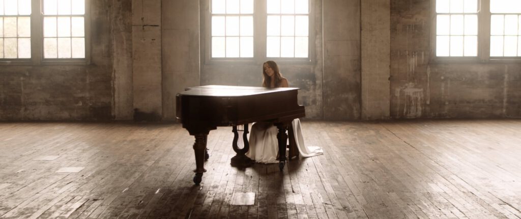 A woman in a long white dress sits at a grand piano in a large, empty room with worn wooden floors and tall windows letting in soft natural light. The atmosphere is serene and slightly nostalgic.