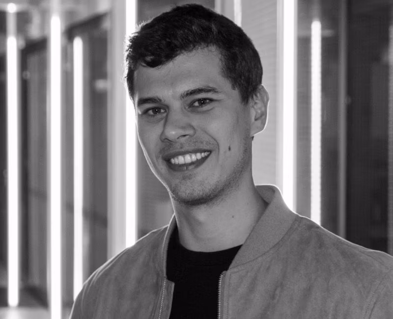 Black and white photo of a young man with short dark hair, smiling at the camera. He is wearing a light-coloured jacket over a dark shirt. Vertical light strips and glass panels form a modern, blurred background.